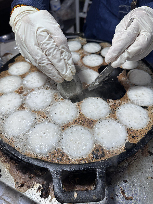 泰国椰子煎饼(Kanom Krok)的特写图像，甜美的街头小吃正在铸铁Ebelskiver风格的平底锅中制作，厨师戴着塑料手套，用金属勺子取出煎饼，高架视图，重点放在前景